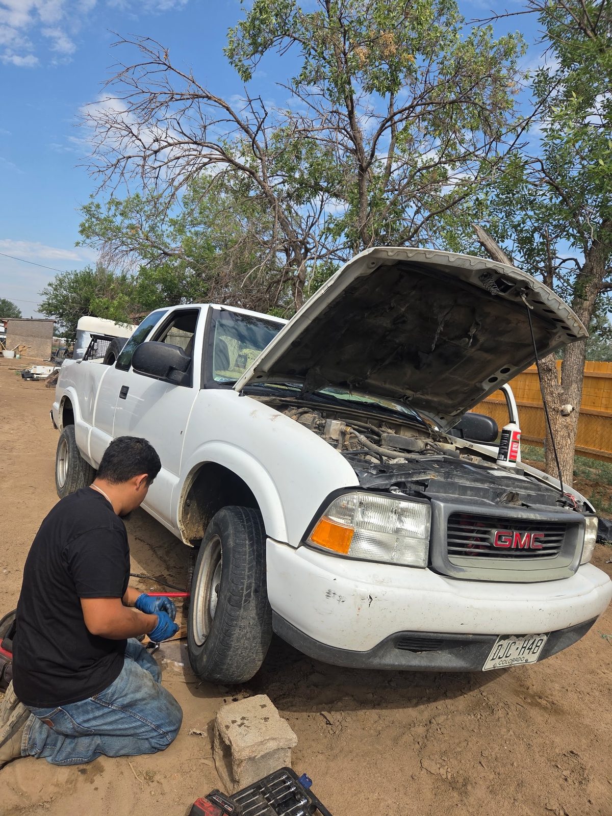 Technician kneeling at the front wheel of a white GMC S10 pickup with the hood up